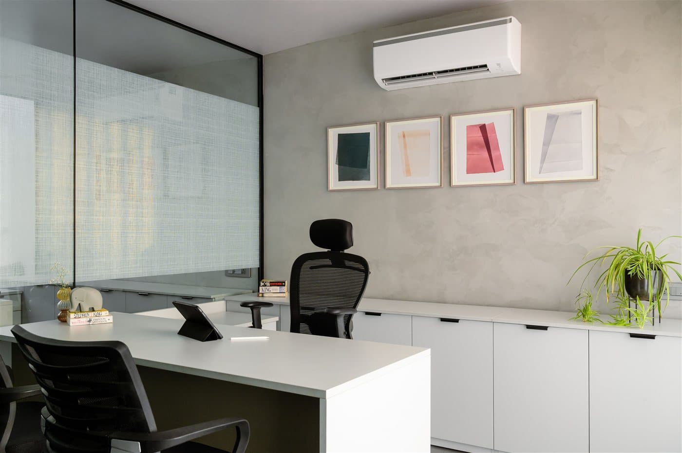 Ruhiges Einzelbüro mit weißem Schreibtisch, Sideboard, Glaswand und gerahmten Bildern an der Betonoptik-Wand